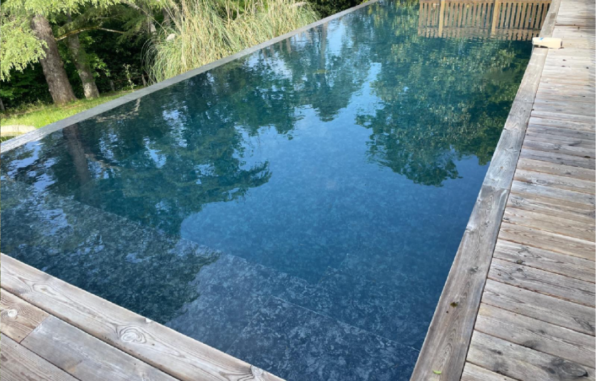 Une vue en perspective d'une élégante piscine à débordement avec finition intérieure en pierre bleu-vert foncé et plage en bois vieilli, réalisée par Couleur Piscine Sud Ouest, expert piscine Arcachon pour des projets haut de gamme. La piscine reflète les arbres environnants dans un cadre naturel préservé.