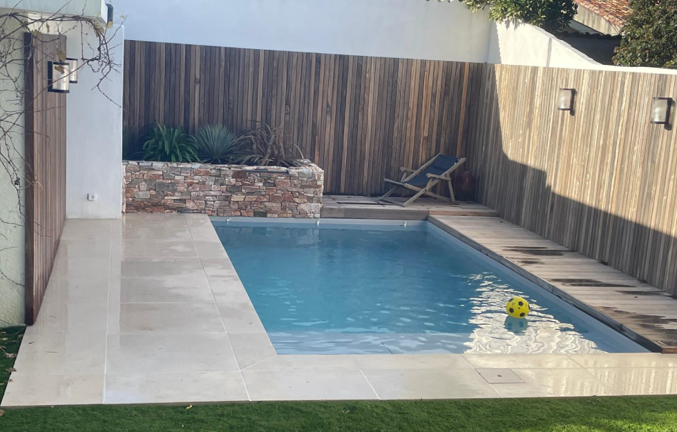 Aménagement d'une piscine citadine Saint-Aubin-de-Carcans dans une cour intimiste, mêlant de larges margelles en pierre claire, une plage en bois et une clôture en claire-voie, conçue par Couleur Piscine Sud Ouest.