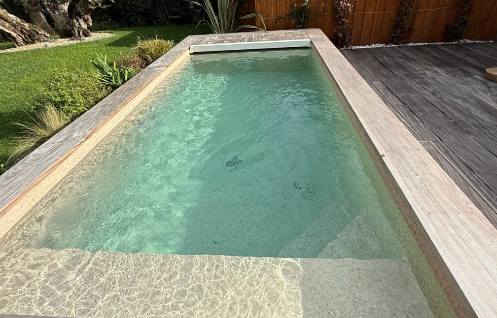 Élégante piscine citadine Lacanau surélevée avec finition lisse grise et larges marches d'accès, posée sur une terrasse en bois au cœur d'une végétation luxuriante, par Couleur Piscine Sud Ouest.