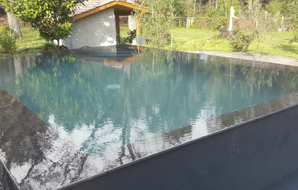 Piscine miroir contemporaine surélevée avec revêtement sombre et margelles en pierre noire, parfaitement intégrée dans un jardin verdoyant par Couleur Piscine Sud Ouest, Expert piscine Lacanau.