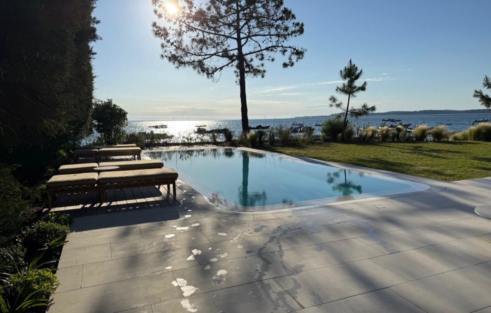 Superbe piscine miroir reflétant un pin majestueux sous le soleil, avec terrasse en pierre claire et bains de soleil face au plan d'eau, une réalisation signée Couleur Piscine Sud Ouest, Expert piscine Lacanau.